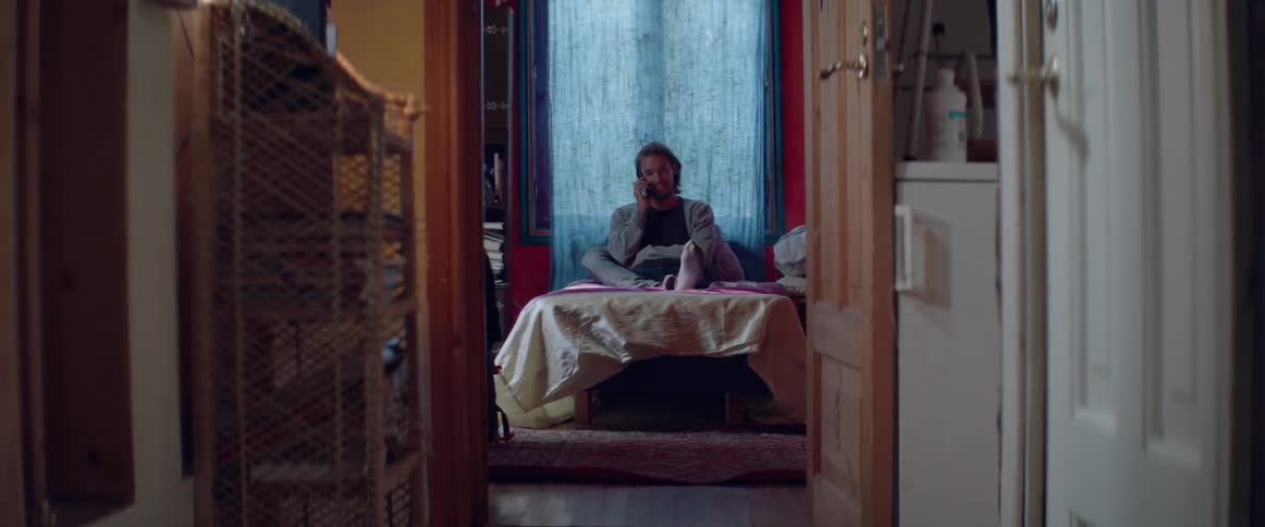 A man is sitting on his bed talking on the telephone during the day