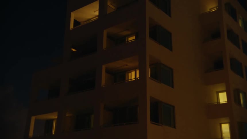 The tall building has balconies with lights on inside