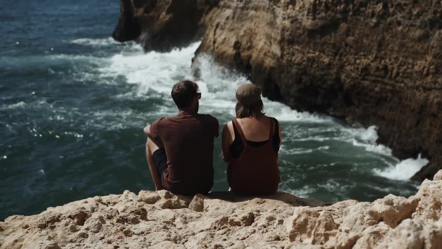 A man and woman sit on a rocky beach