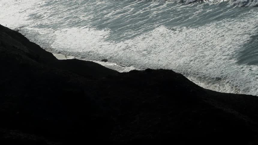 Waves are rolling toward a beach at the base of cliffs during the afternoon