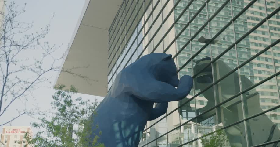 Large statue of a blue bear leans against a building with many glass windows