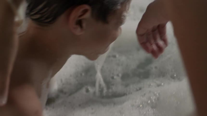 A boy in a bathtub playing with a shark before he puts his face under the water, lifts back up and a woman brushes the bubbles away from his eyes