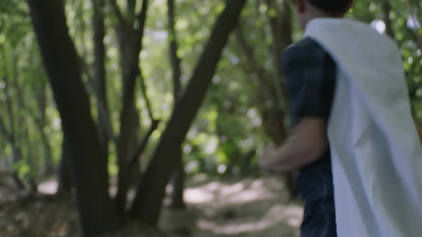 A boy runs through the forest with a piece of cloth worn as a cape