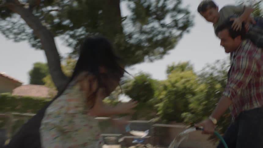 Man chases a child around a yard as a girl sprays water on them