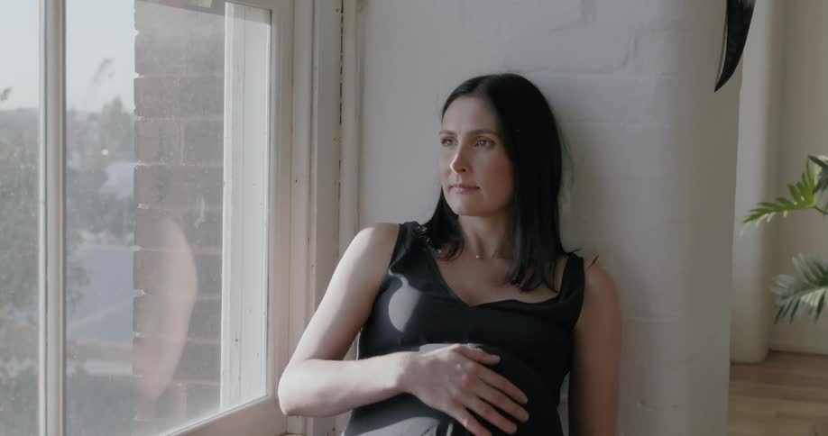 A pregnant woman sits on the window looking outside