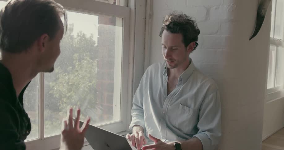 Two men sit on a window ledge with a laptop and talk to each other