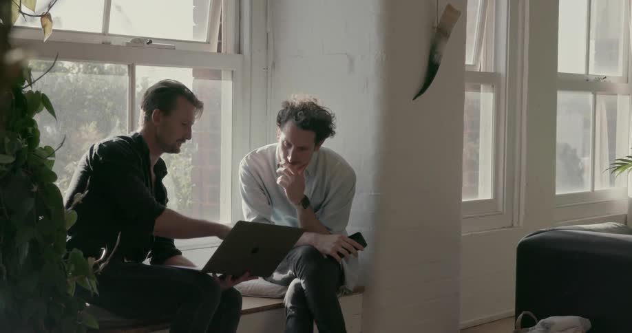Two men sit near a window looking at a laptop
