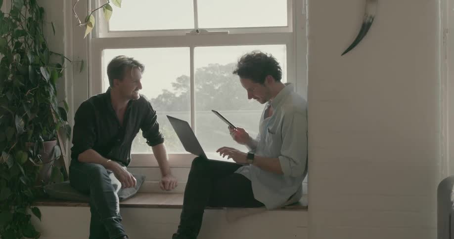 Two men sit on a window ledge with a laptop and cell phone