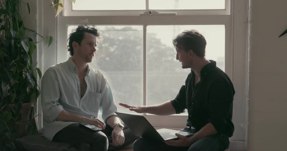 Man with a laptop talks to another man while sitting near a window