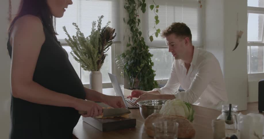 A woman cuts food as a man uses a laptop