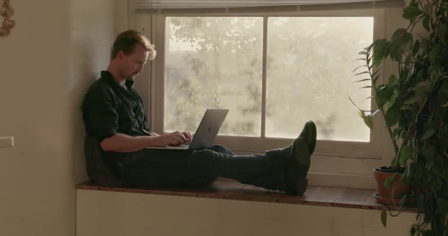 Man sits in a window sill typing on a laptop
