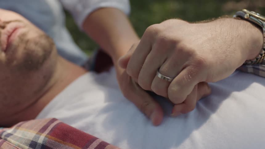 A man and a woman clasp hands on the chest of a man lying down, and he then clasps their hands