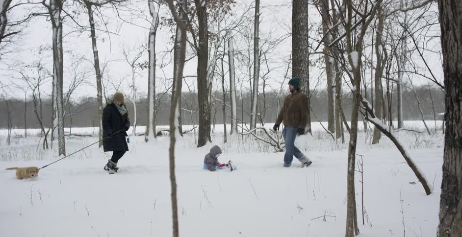 A woman walks a dog as a man pulls a child on a sled