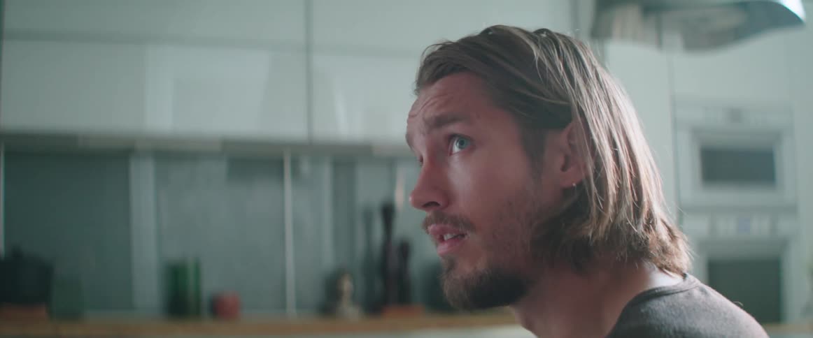 A confused young man sitting at a kitchen table looks up while talking.