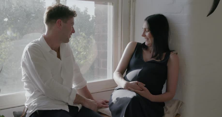 Man and pregnant woman sitting nest to a window as she leans on the wall and laughs