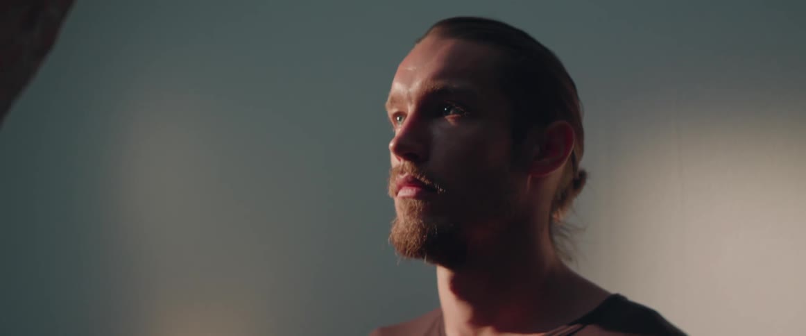 A serious bearded man blinks slowly while standing inside a dimly lit room.