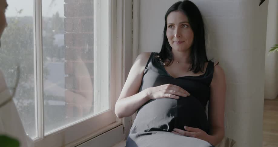 A pregnant woman holds her belly by the window