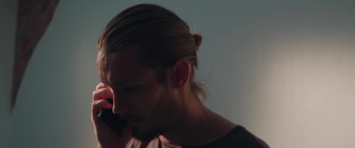 A frustrated male character stands under dim lighting having a disappointing phone conversation.