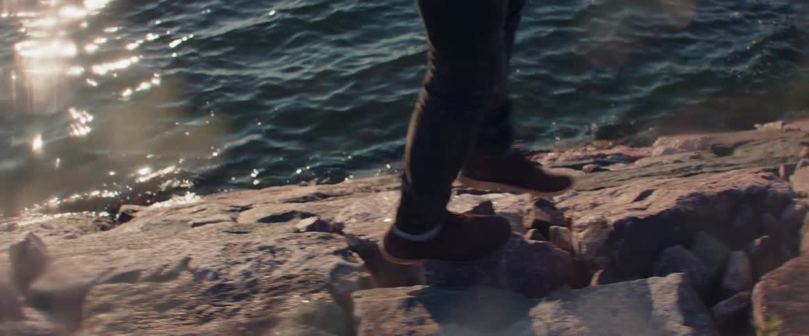 Low angle shot of a person’s feet walking on rocky shoreline near water.