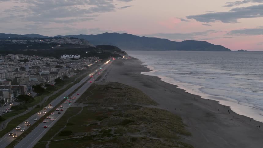 Vehicles are driving in controlled groups down a coastal highway at dusk