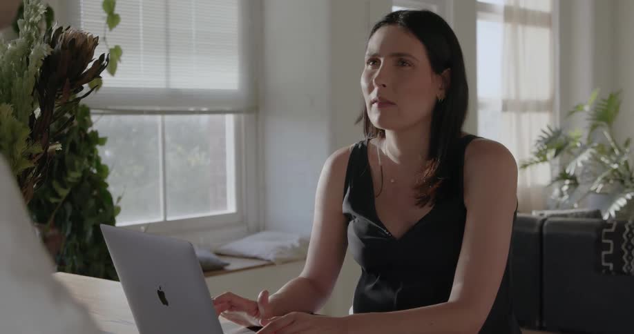 A pregnant woman sits at the counter talking and typing on a laptop