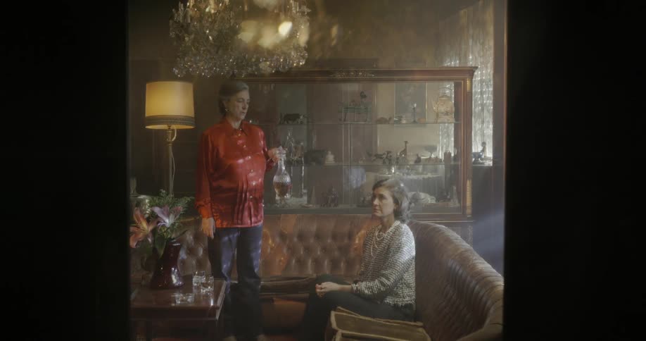 Woman lifts a decanter as she stands near a sofa where another woman sits