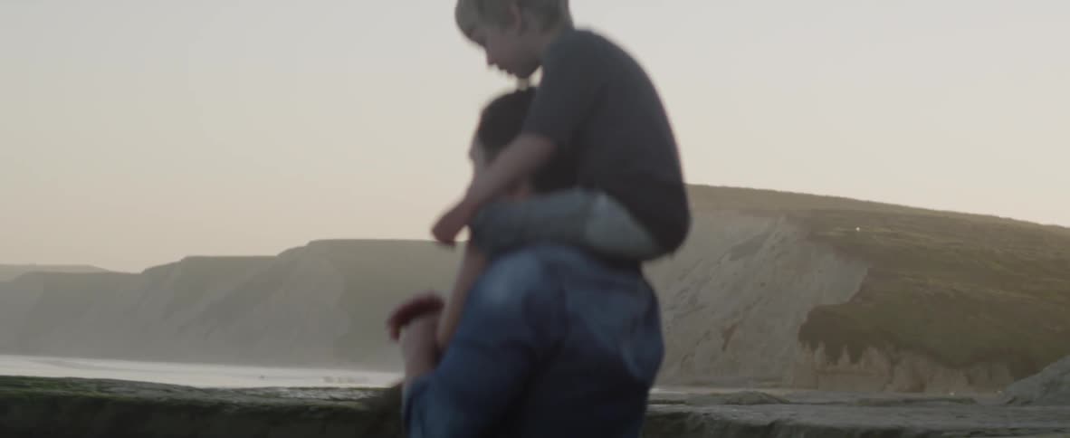 A father and son stand together on a rugged beach, looking toward the horizon at sunset