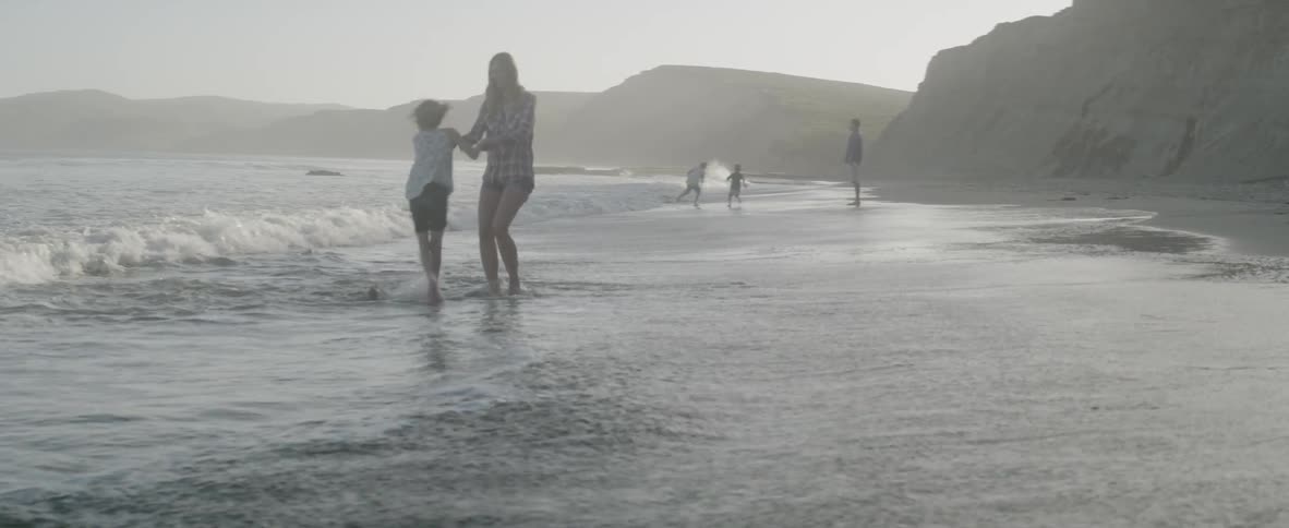 A mother and daughter spin together at the shoreline as their family plays nearby