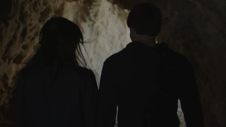 A couple holds hands as they exit a large cave with a sandy floor