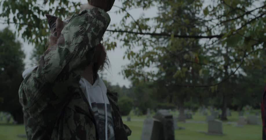 A teenager wearing a mask is walking through a cemetery and drinking at dusk