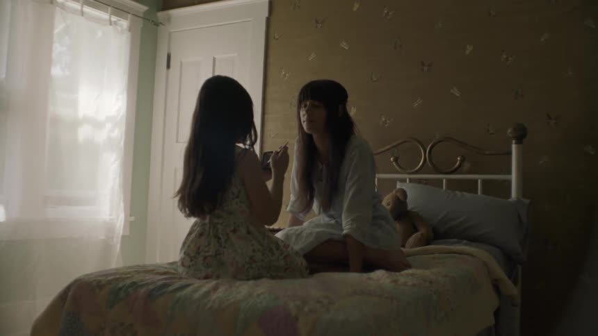 Two girls kneel on a bed while one applies makeup to the other
