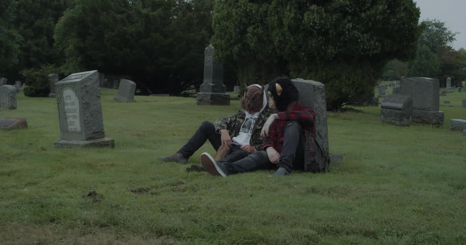 Two people wearing masks sit against a gravestone talking