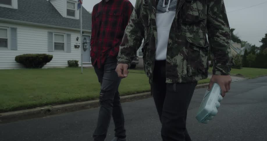 Two men walk down a neighborhood street while one carries a carton of eggs