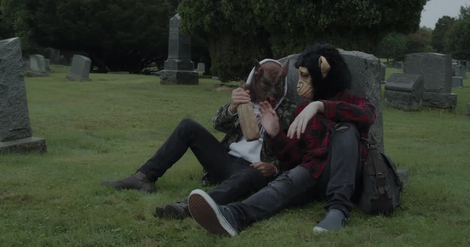 Two people in masks sit against a tombstone drinking