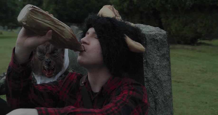 Boys wearing masks lean against a headstone and drink from a bottle in a paper bag