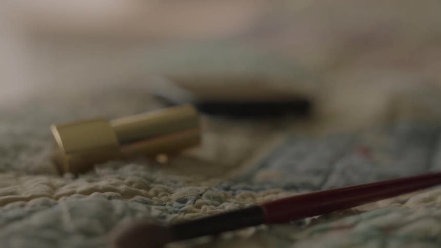A compact, lipstick tube and makeup brush laying on a quilt