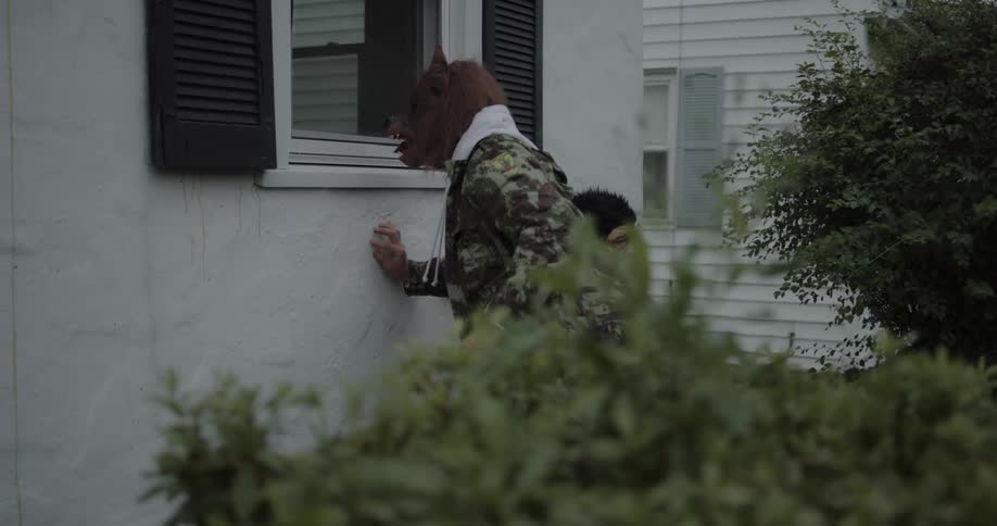 Two people wearing animal masks are looking in window of house