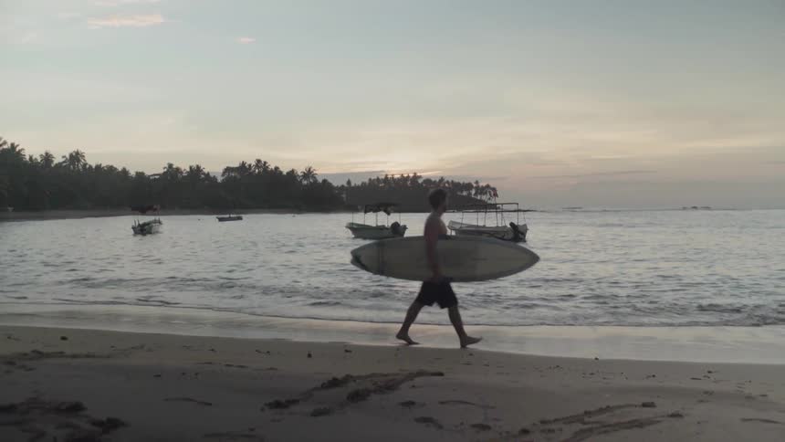 Man is walking on beach while carrying surfboard at sunset