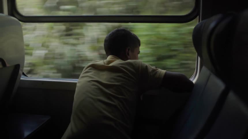 A boy is leaning out of a train window watching the scenery roll by in daylight