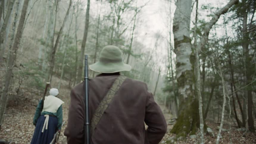 A man and woman carry rifles through the woods