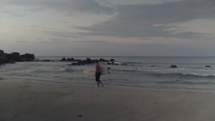 A surfer is carrying a surfboard near the water's edge of a beach at dusk