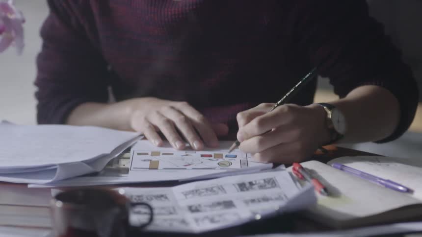 A man is writing on paperwork as he sits at a table inside in daylight