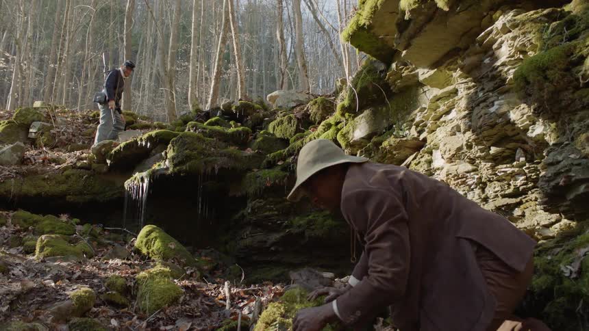 A man searches around in rocks and leaves as a soldier stands watch