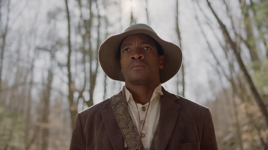 A black man is standing in a natural setting staring straight ahead in daylight