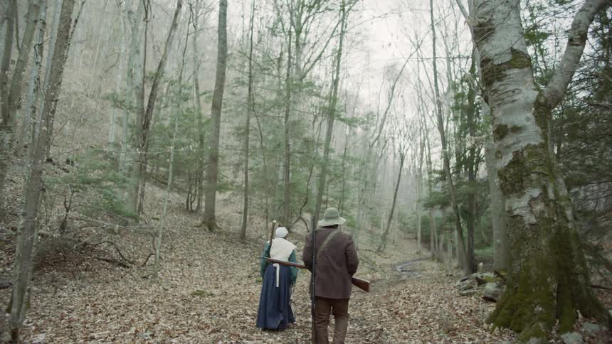 People walk through a forest with rifles and 19th century clothing