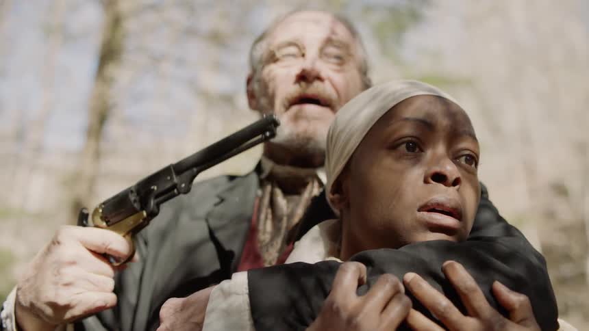 A man is shooting off a gun and threatening a slave woman as they stand outside in daylight