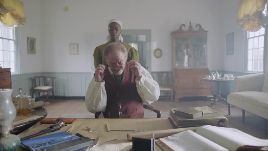 Man leans on a desk writing as a woman carrying a teacup walks up behind