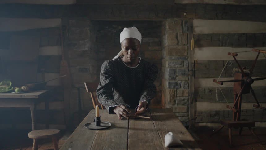 A slave woman is picking up a pipe from a table inside a cabin in daylight