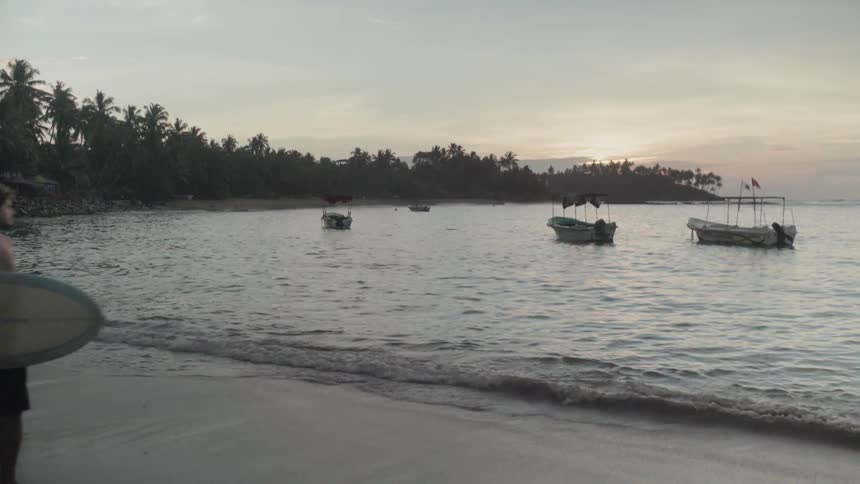 A surfer walks by anchored boats on the beach