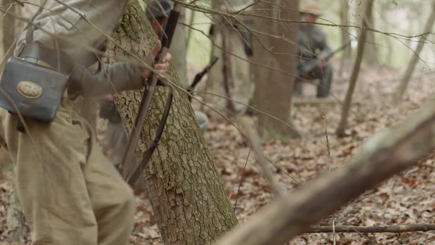Confederate soldiers are firing their rifles as they move through a forest in daylight